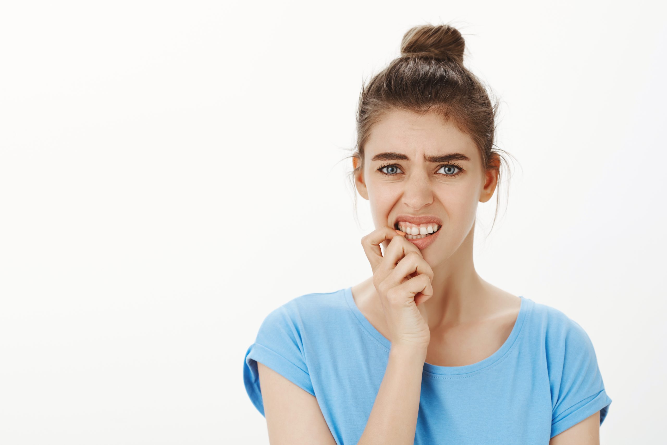 Waist-up shot of intense worried european woman with bun hairstyle, frowning, clenching teeth and biting fingernail anxiously, standing nervous over gray background, having terrible paranoia